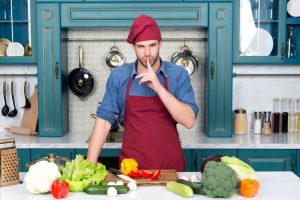 Chef standing in his kitchen, with ingredients out to prepare a dish, but he has a finger to his lips because he will not give away his secret ingredient.