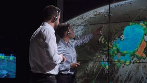 A man and a woman, who are both wearing headsets, are looking at a large-screen weather map