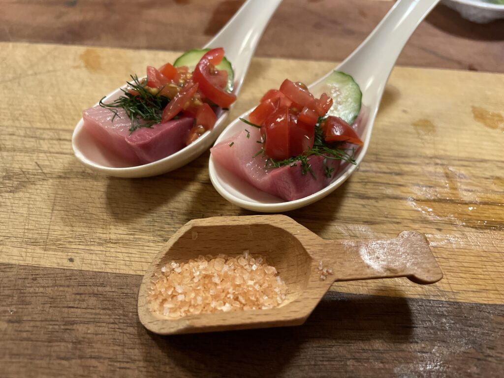 Two spoons with pink fish, tomato and seasoning with one wood scoop holding pink sea salt