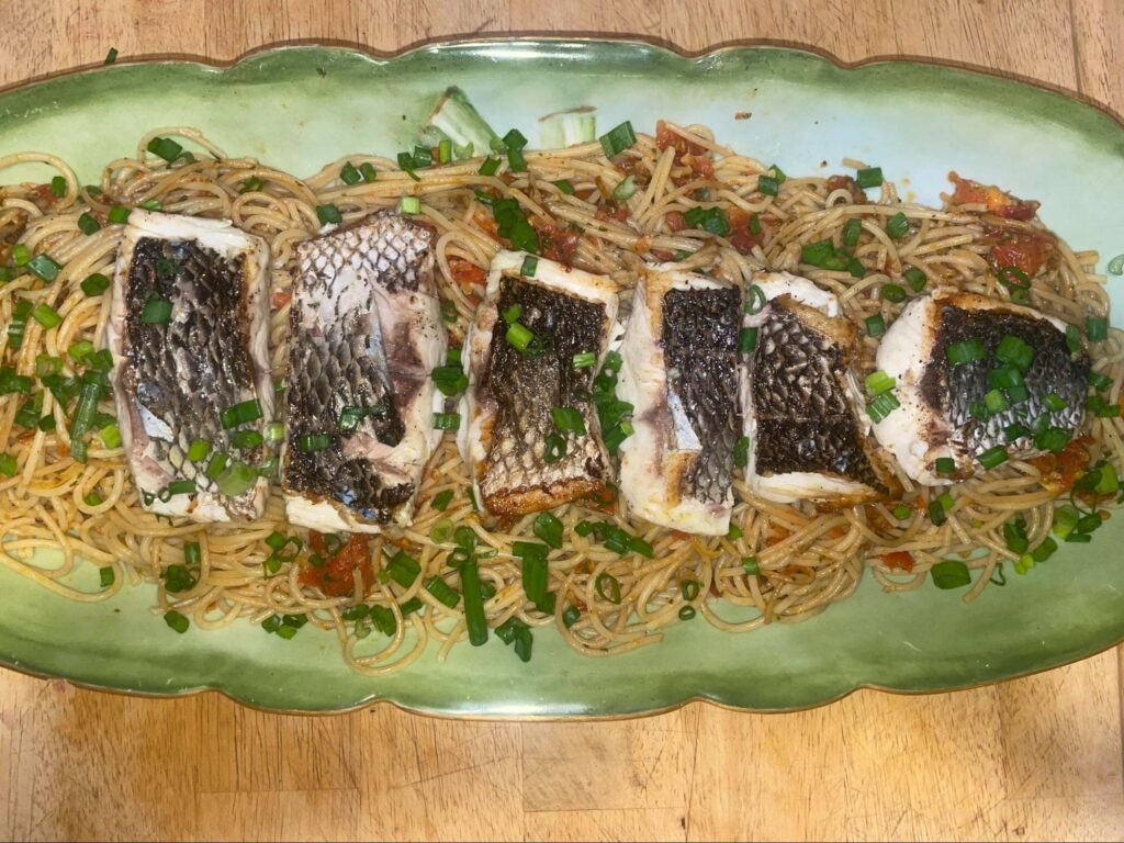 A green platter on a tan tabletop with pasta noodles, green basil, red tomatoes, and a silver, shiny skin on four fillets of a white-fleshed fish.