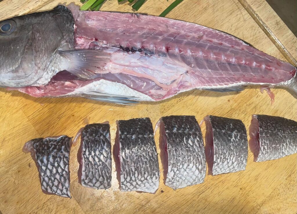 Silver fish on tan cutting board with six filets portioned out of the side of the fish, laying beside the rest of the body of the fish. The fish meat is a mixture of silver, white and purple shades ombre throughout the meat.